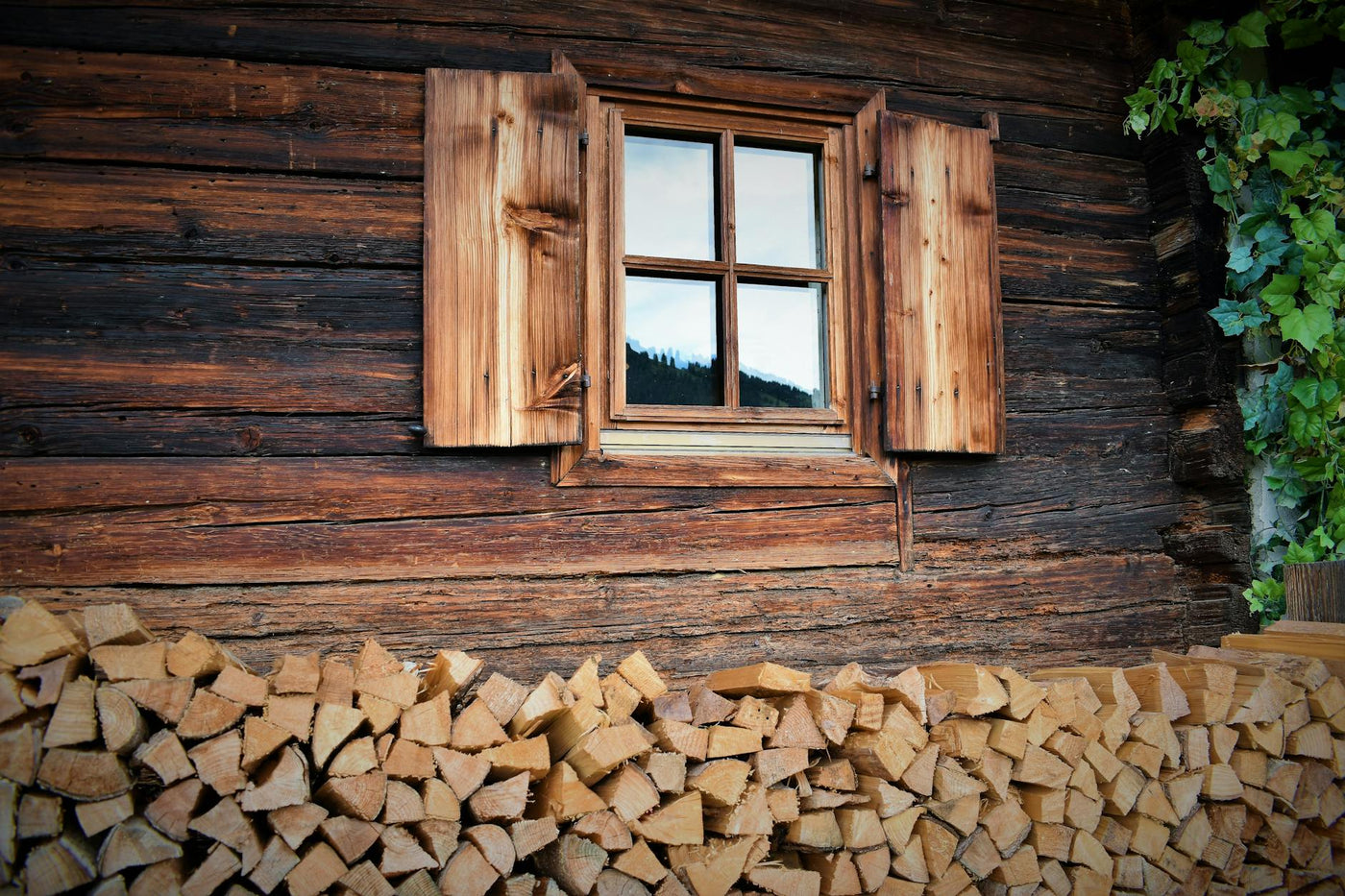 Rustic cabin wall with wooden windows and neatly stacked logs, reflecting serene mountain scenery.