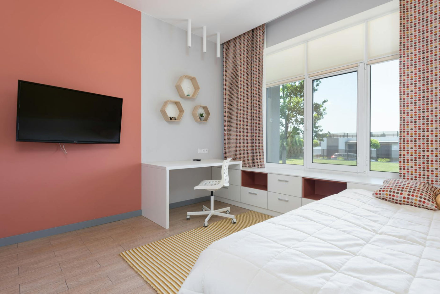 Modern design of comfortable bedroom with light workplace near big window and wide bed in opposite of TV set on coral wall