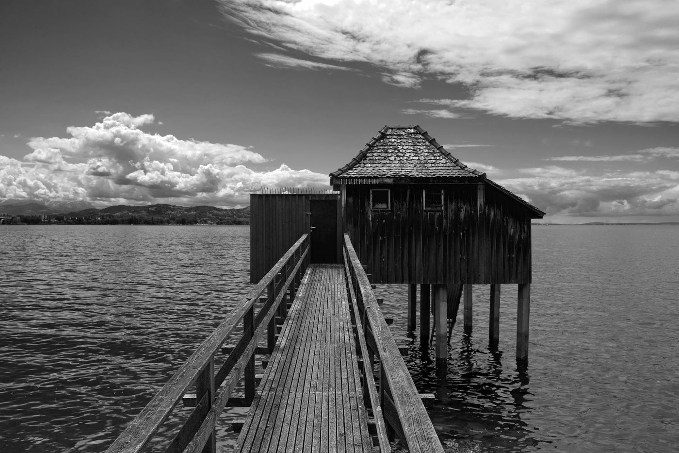 Dramatic black and white photo of a wooden pier leading to a hut over a lake under a cloudy sky.